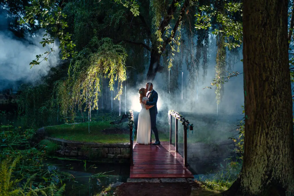 Ein Paar umarmt sich auf einer kleinen Holzbrücke, umgeben von üppigem Grün und Nebel. Licht fällt durch die Blätter und schafft eine magische und romantische Atmosphäre, perfekt eingefangen von Hochzeitsfotograf Roman Koryzna.