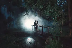 Ein Paar steht auf einer Holzbrücke in einem nebligen Wald, umgeben von Bäumen und sanftem Licht.