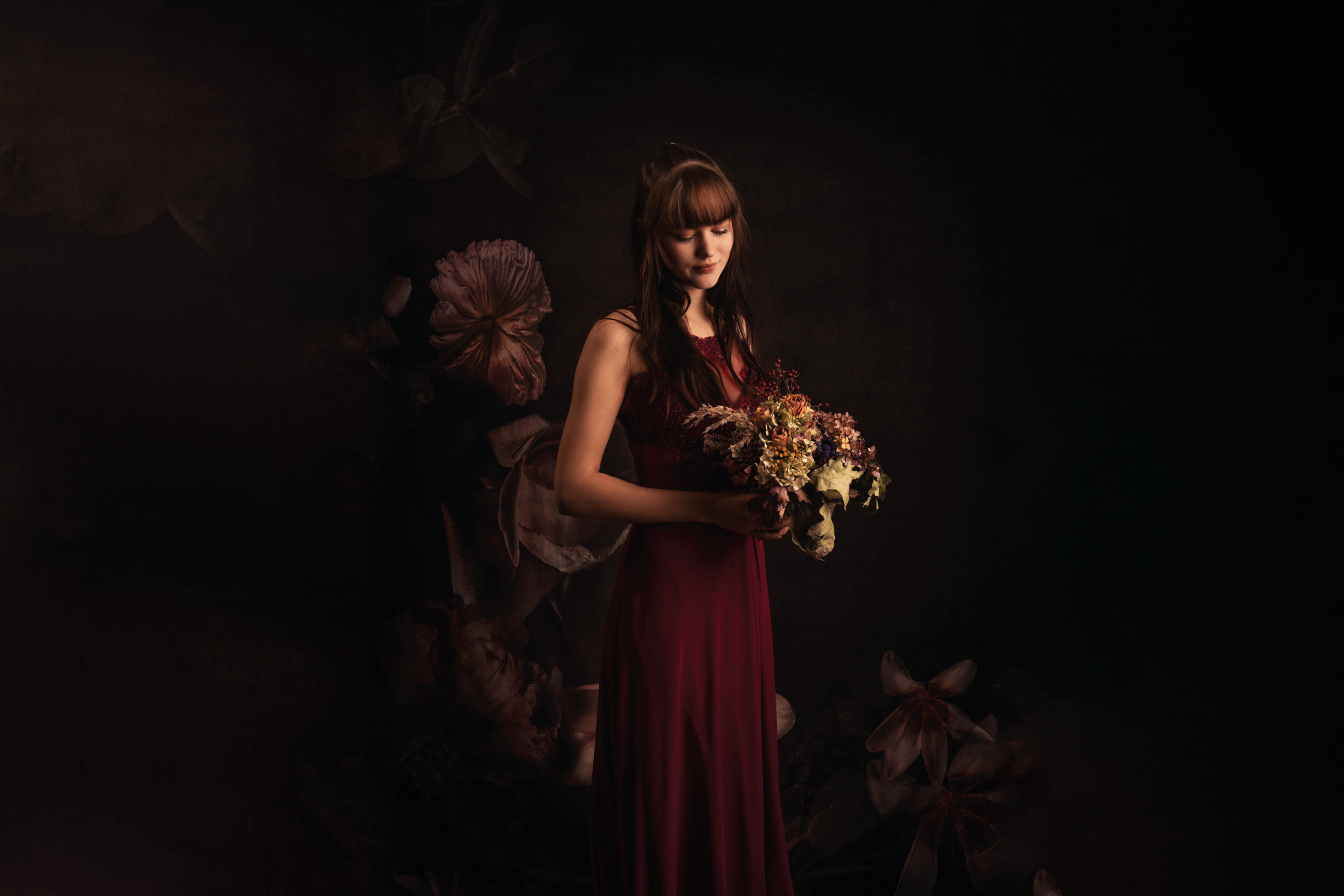Eine Frau in einem dunkelroten Kleid, aufgenommen vom Fotografen Roman Koryzna, hält einen Blumenstrauß in der Hand. Sie steht vor einem dunklen, floralen Hintergrund. Ihr Blick ist nach unten gerichtet, was eine heitere und besinnliche Atmosphäre schafft.