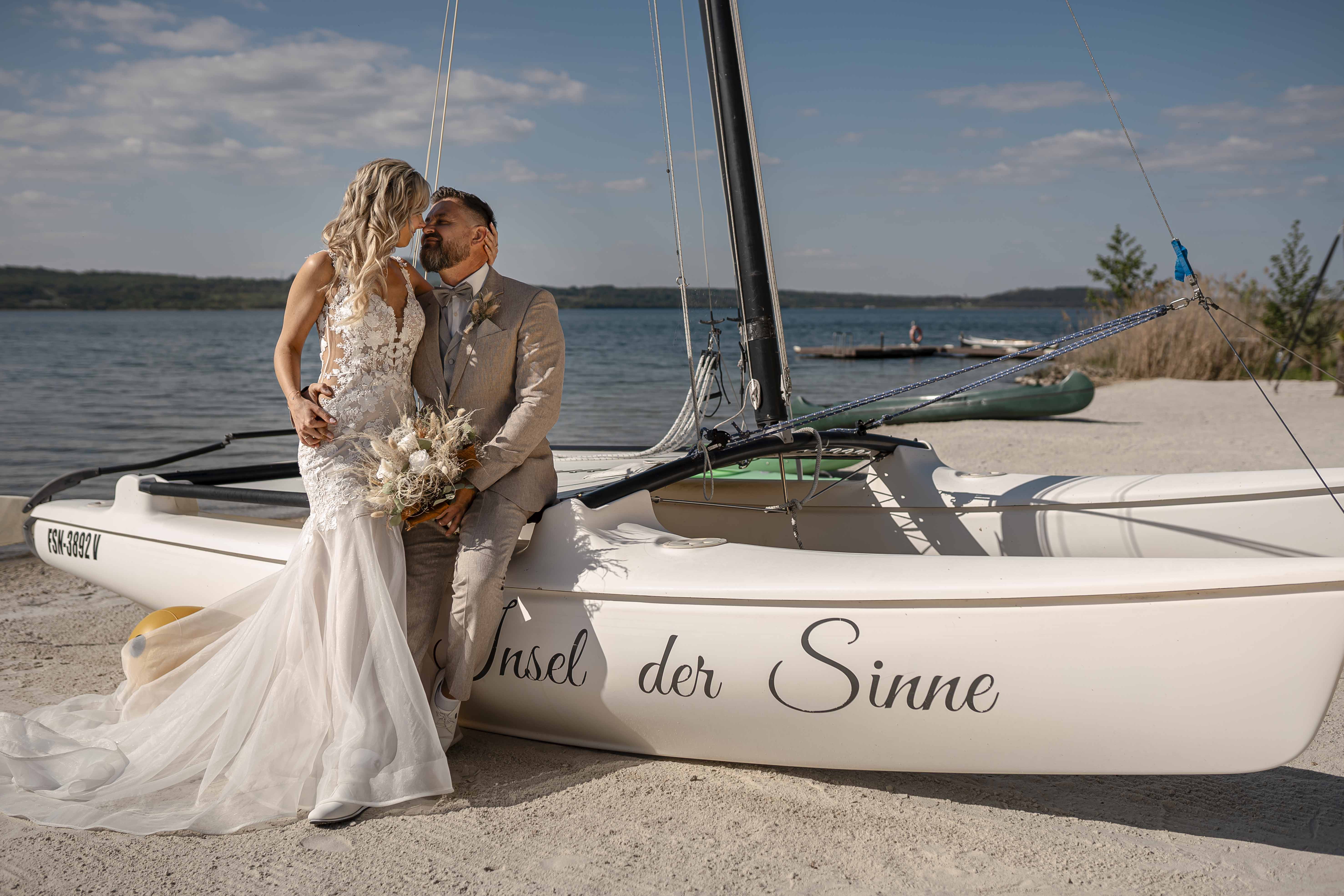 Braut und Bräutigam posieren an einem Hochzeitsboot am Wasser, umgeben von einer romantischen Kulisse mit Blumenstrauß.