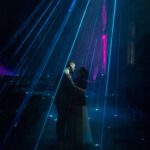 Ein Paar umarmt sich unter neonblauen und pinken Lichtern, ihre Silhouetten heben sich dramatisch vom dunklen Hintergrund ab. Laserstrahlen bilden einen Baldachin um sie herum und verbreiten eine romantische und himmlische Atmosphäre – perfekt eingefangen von Hochzeitsfotograf Roman Koryzna.