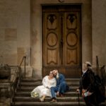 Ein Paar in formeller Kleidung, die Frau im weißen Kleid und der Mann im blauen Anzug, sitzt glücklich auf Steinstufen. Sie posieren liebevoll vor einer großen, verzierten Holztür, während Hochzeitsfotograf Roman Koryzna den Moment einfängt, während eine Person mit einem Spazierstock vorbeigeht.