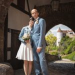 Hochzeitspaar posiert unter einem historischen Bogen, Braut mit Blumenstrauß und Bräutigam in Anzug, romantische Atmosphäre. Foto: Roman Koryzna