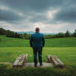 Ein Mann in einem blaugrünen Anzug steht mit dem Rücken zur Kamera und blickt über eine üppig grüne Landschaft unter einem bewölkten Himmel. Aufgenommen von Hochzeitsfotograf Roman Koryzna steht er auf einem Steinpfad, in dessen Ferne sich Grasfelder erstrecken.