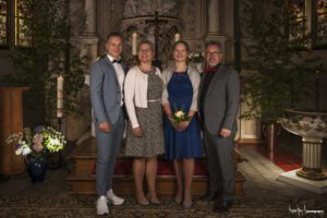 Konfirmation in der Kirche von Milkel - Roman Koryzna Fotografie | Der Hochzeitsfotograf für Bautzen und Dresden Eine Gruppe von vier Personen steht in einer mit Kerzen und Blumenarrangements geschmückten Kirche. Die Frauen, die Blumen halten, tragen Abendkleider, während die Männer Anzüge tragen. Die Konfirmationsfotografie, aufgenommen mit der Linse von Roman Koryzna, hebt die elegante Umgebung mit aufwendigen Dekorationen und Buntglas hervor.
