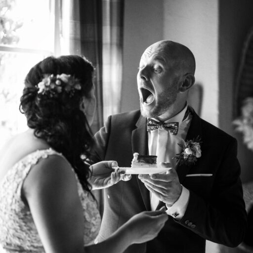 Ein Brautpaar in festlicher Kleidung, der Bräutigam zeigt Überraschung, während er ein Stück Kuchen von der Braut erhält. Hochzeitsfeier-Moment. Foto: Roman Koryzna