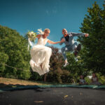 Braut und Bräutigam springen gemeinsam auf einem Trampolin in einem Garten. Foto: © Roman Koryzna, Hochzeitsfotograf. Weißenberg 2022. Hochzeitsfotograf für Bautzen Dresden und die Lausitz.