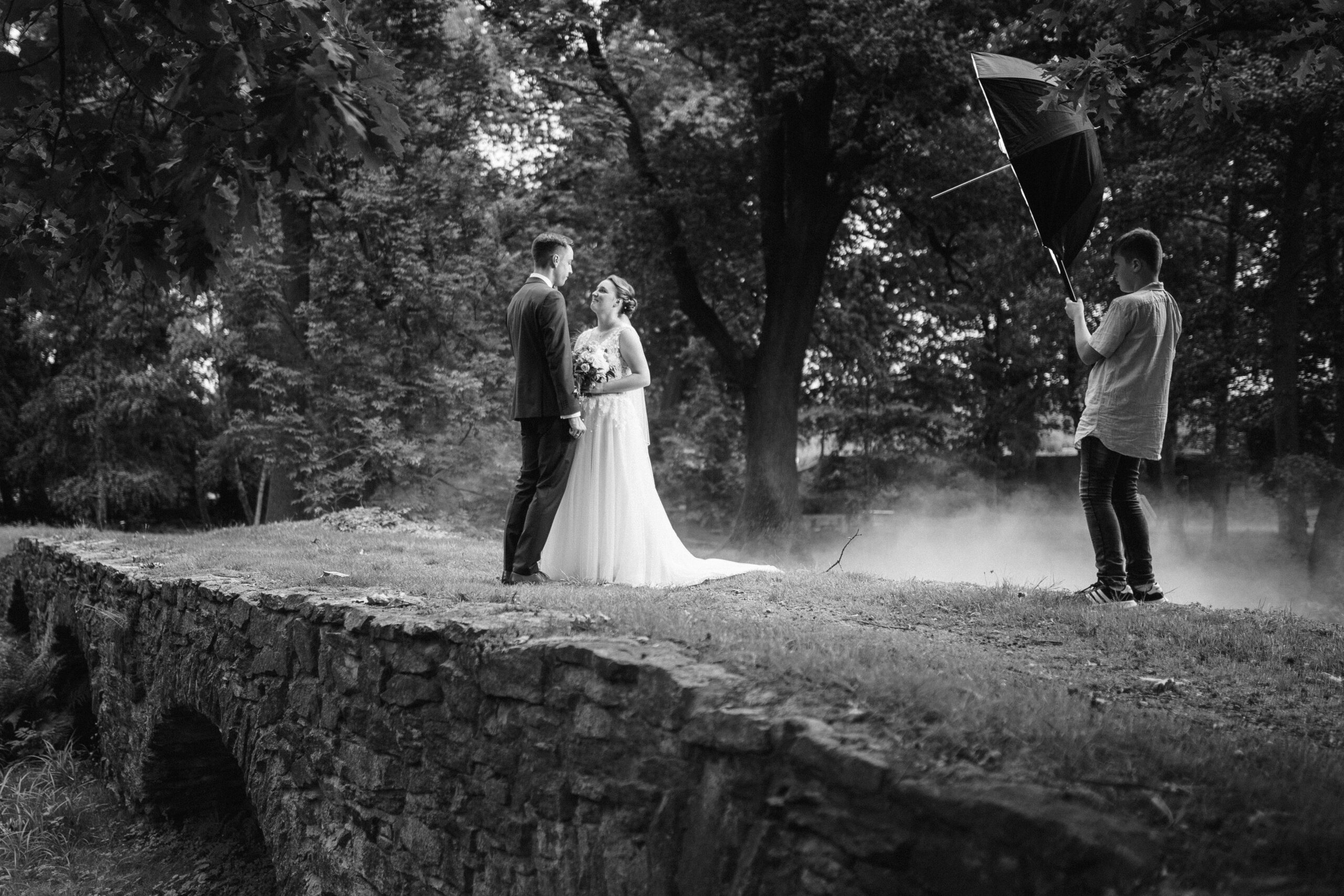 Hochzeitsfotografie von Celine und Philipp Ein Brautpaar steht auf einem Steinweg während eines Fotoshootings mit einem Blitzschirm. Foto: © Roman Koryzna 2023. Hochzeitsfotograf für Bautzen Dresden und die Lausitz.