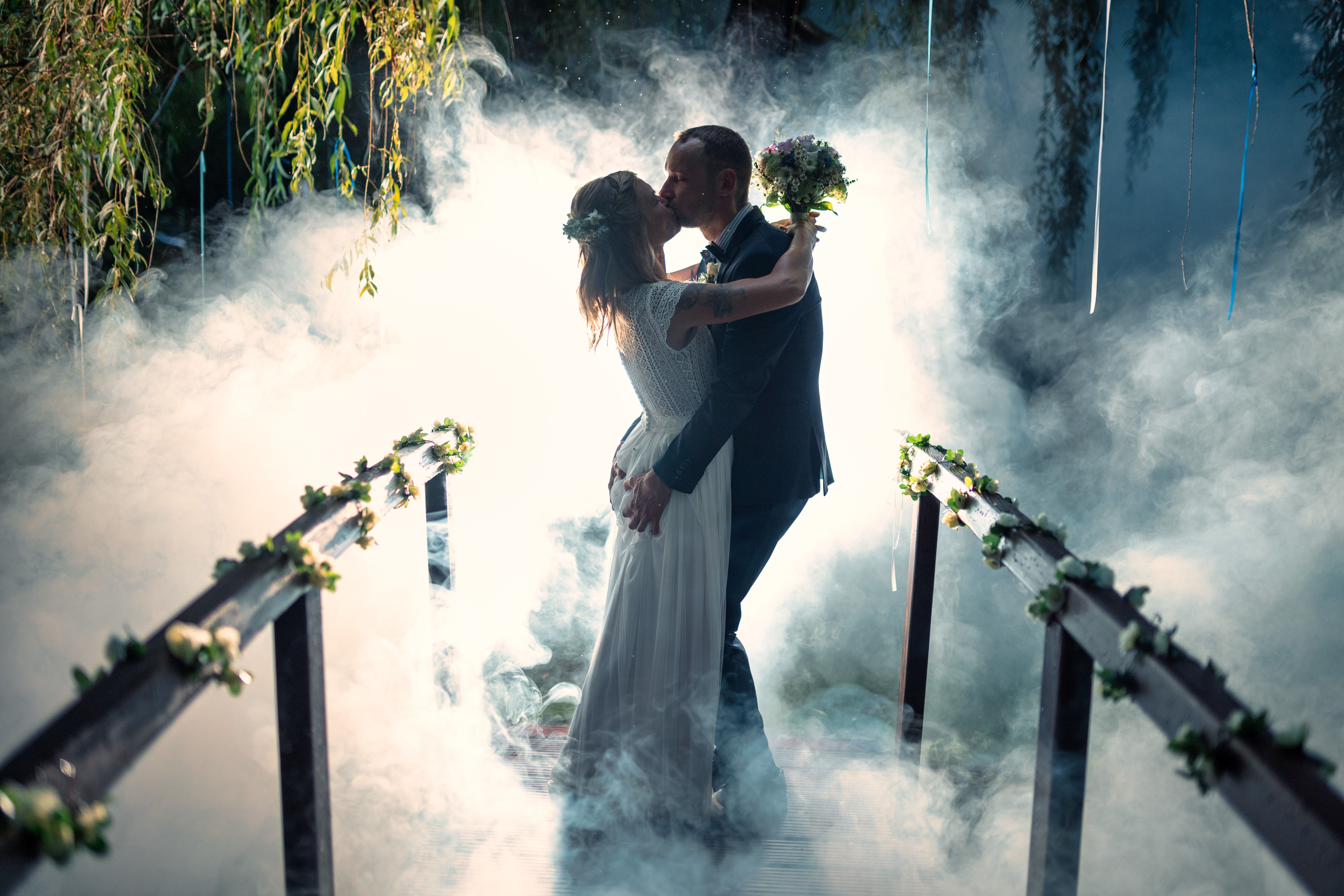 Ein Brautpaar küsst sich auf einer Brücke umgeben von Nebel und Blumenarrangements. Foto: © Hochzeitsfotograf Roman Koryzna, August 2022. Hochzeitsfotograf für Bautzen und Dresden