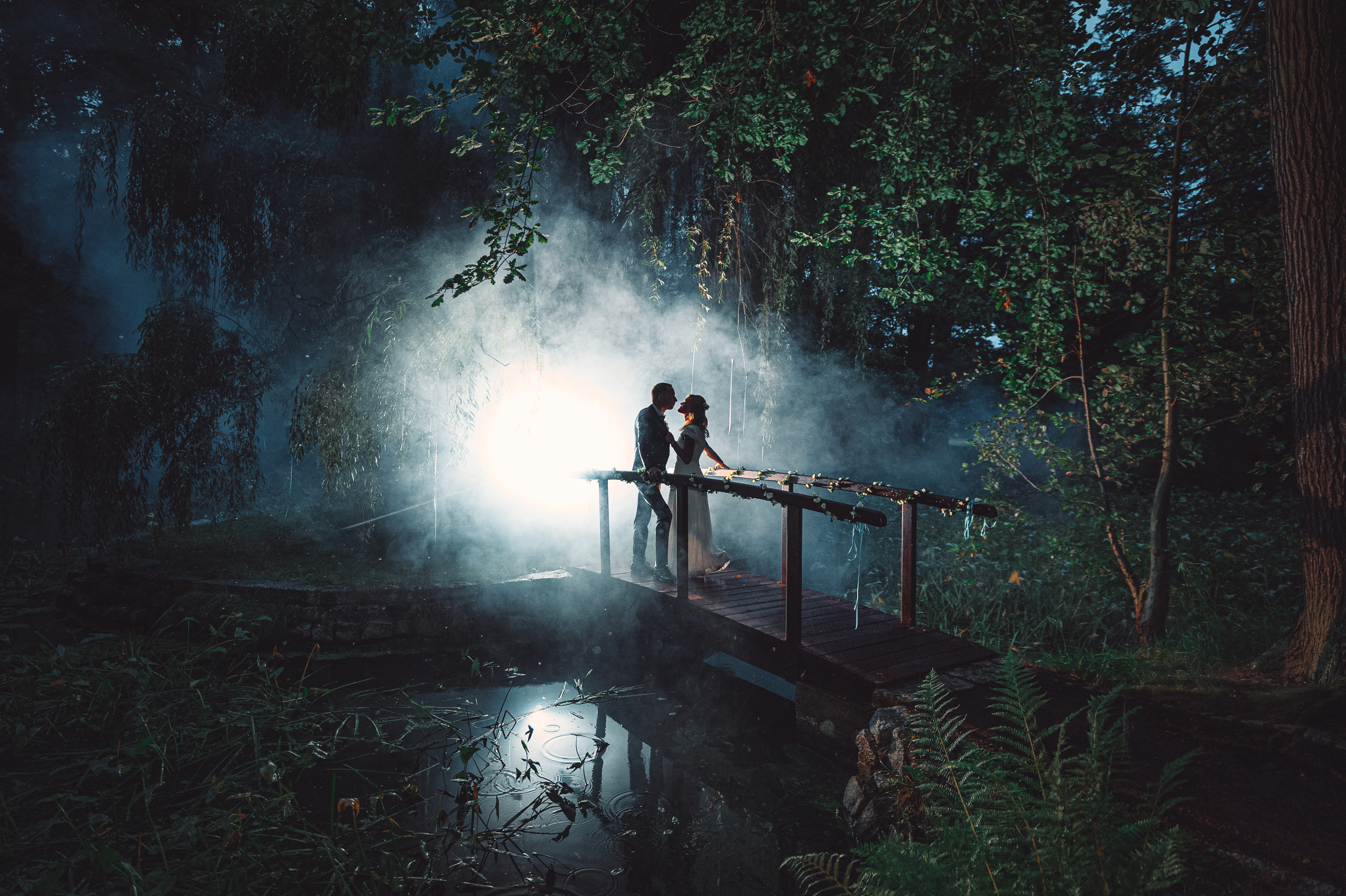 Paar steht auf einer Brücke in einem nebligen Wald mit Licht im Hintergrund. Foto: © Hochzeitsfotograf Roman Koryzna, August 2022. Hochzeitsfotograf für Bautzen und Dresden