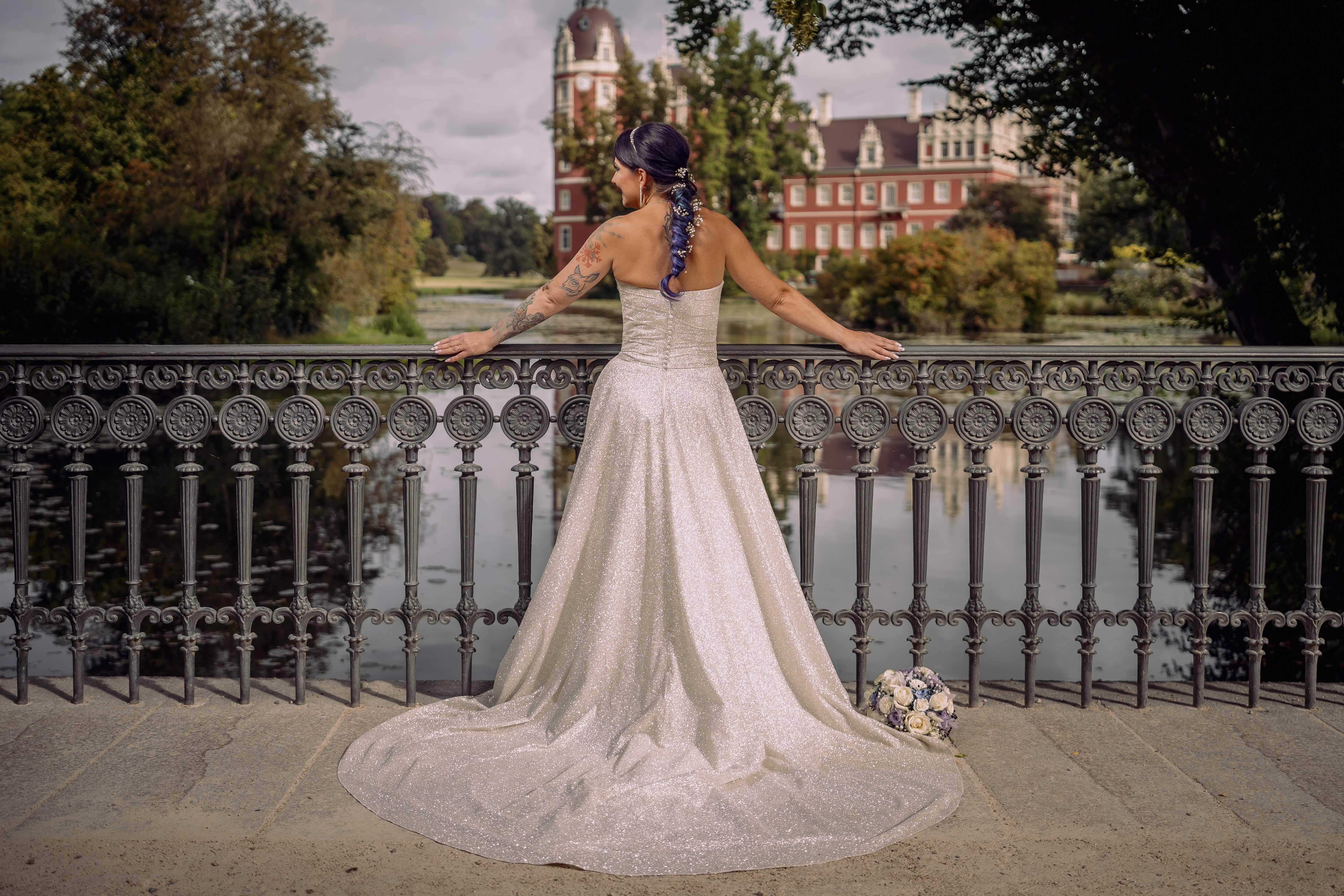 Braut in einem glitzernden Hochzeitskleid steht mit dem Rücken zur Kamera an einem Geländer. Fürst-Pückler-Park in Bad Muskau. © Foto: Hochzeitsfotograf Roman Koryzna
