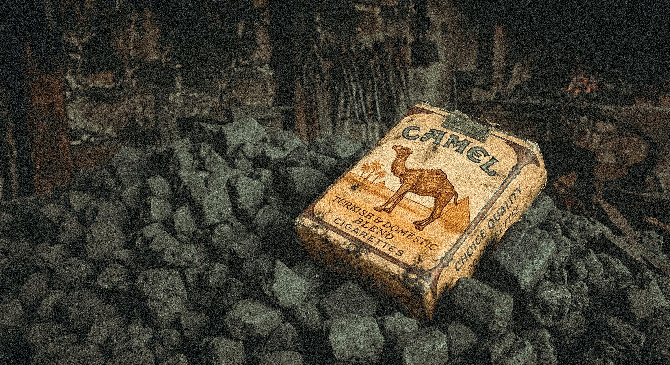 Discarded Camel cigarette pack lying on a bed of dark rocks in a dim, smoky setting with a small fire in the background.