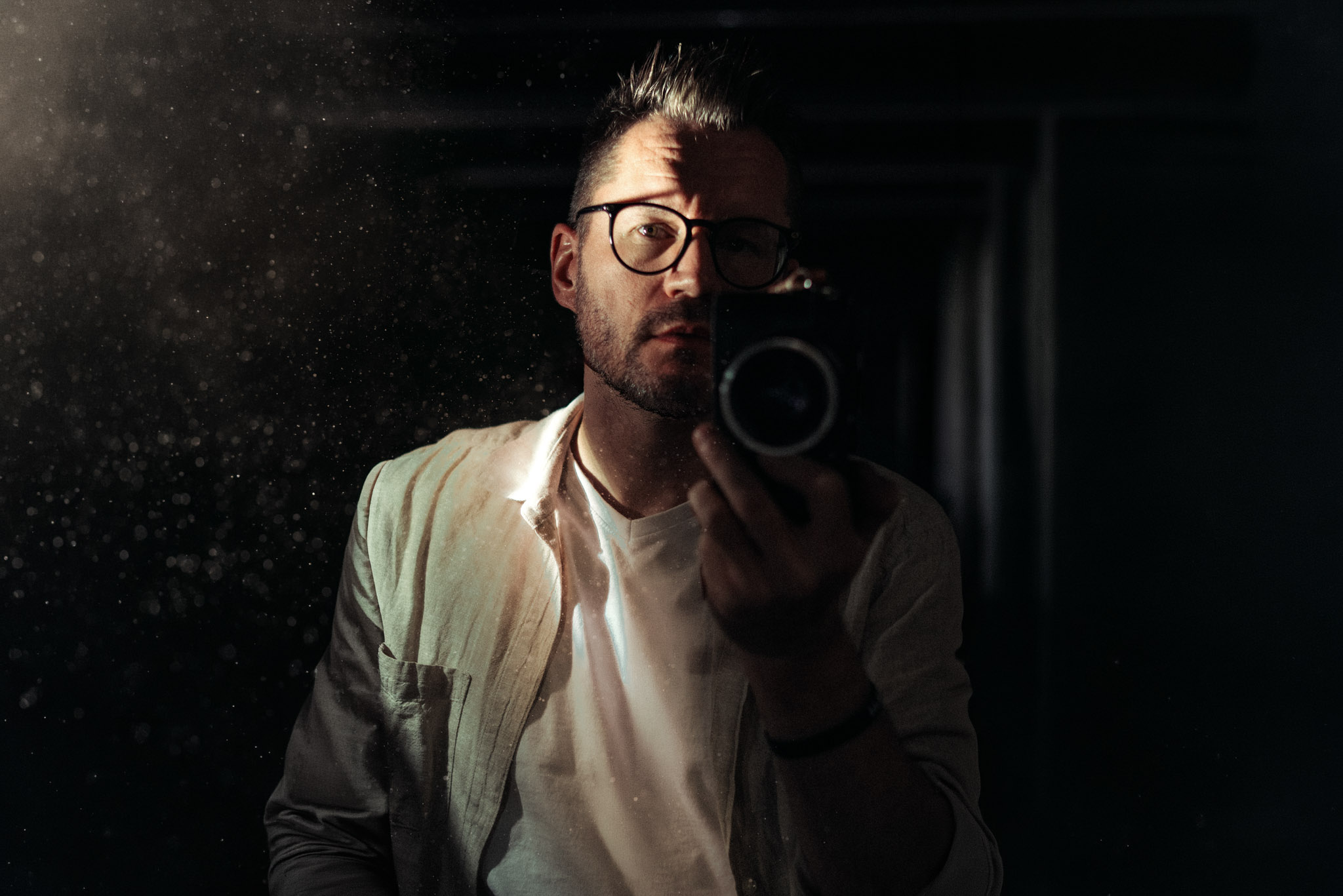 Man wearing glasses takes a mirror selfie in a dark, dusty room with light specks in the air; camera partly covers his face.