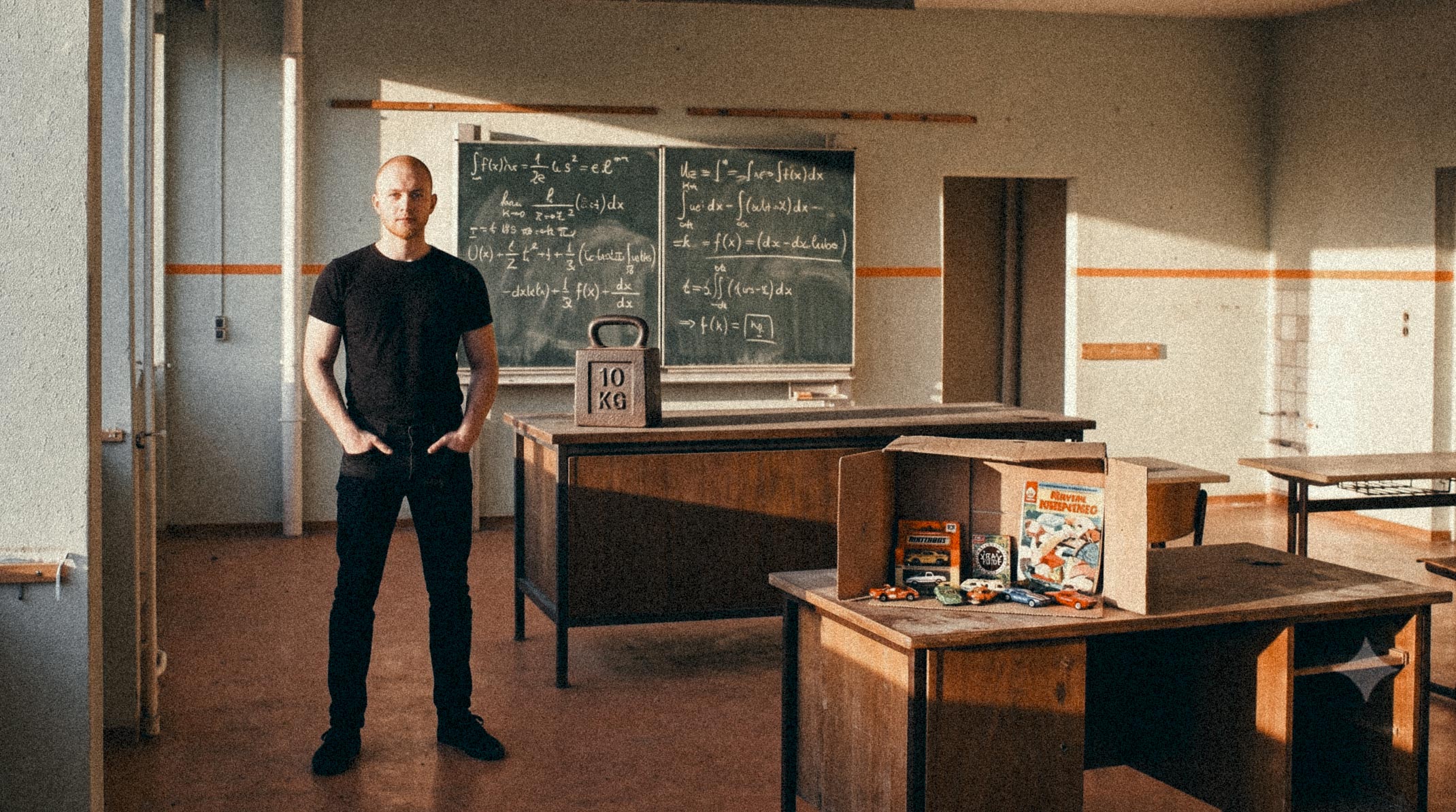 Man in a black t-shirt stands in a classroom or lab, with chalkboards full of equations behind him and a 10 kg weight on a desk nearby; toy boxes and small cars sit on a table to the right.